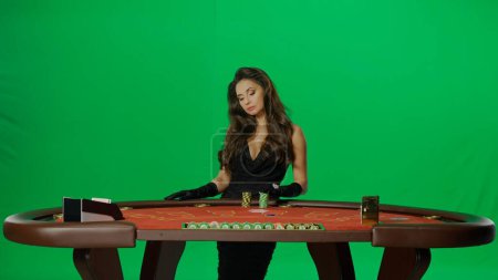 Photo for Casino and gambling commercial advertisement concept. Elegant female in studio on chroma key green screen. Attractive woman in black dress at the blackjack poker table looking at bets. - Royalty Free Image
