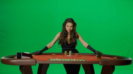 Photo for Casino and gambling commercial advertisement concept. Elegant female in studio on chroma key green screen. Attractive woman in black dress at the blackjack poker table looking at camera. - Royalty Free Image