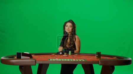 Téléchargez les photos : Casino et jeu publicité commerciale concept. Elégante femme en studio sur écran vert chromé. Belle femme en robe noire à la table de poker blackjack posant à la caméra. - en image libre de droit