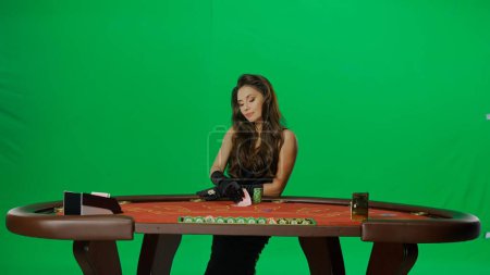 Photo for Casino and gambling commercial advertisement concept. Elegant female in studio on chroma key green screen. Attractive woman in black dress at the blackjack poker table checking cards. - Royalty Free Image