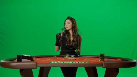 Photo for Casino and gambling commercial advertisement concept. Elegant female in studio on chroma key green screen. Attractive woman in black dress at the blackjack poker table drinking champagne. - Royalty Free Image