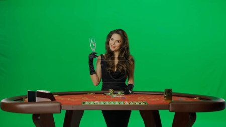 Photo for Casino and gambling commercial advertisement concept. Elegant female in studio on chroma key green screen. Attractive woman in black dress at the blackjack poker table posing with champagne. - Royalty Free Image