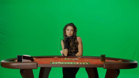 Photo for Casino and gambling commercial advertisement concept. Elegant female in studio on chroma key green screen. Attractive woman in black dress at the blackjack poker table looks at camera. - Royalty Free Image
