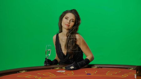 Photo for Casino and gambling commercial advertisement concept. Female in studio on chroma key green screen close up. Woman in black dress at the blackjack poker table holding glass of champagne, posing. - Royalty Free Image