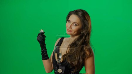 Photo for Casino and gambling commercial advertisement concept. Elegant female in studio on chroma key green screen background. Attractive woman in black dress with champagne and chip looking at camera - Royalty Free Image