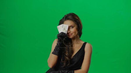 Photo for Casino and gambling commercial advertisement concept. Elegant female in studio on chroma key green screen background. Attractive woman in black dress looking at camera hiding behind ace cards. - Royalty Free Image