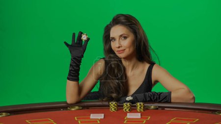 Photo for Casino and gambling commercial advertisement concept. Female in studio on chroma key green screen close up. Woman in black dress sitting at the blackjack poker table smiling holding chip. - Royalty Free Image