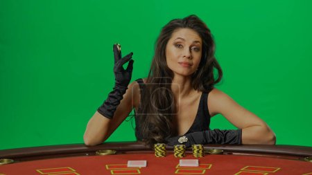 Photo for Casino and gambling commercial advertisement concept. Female in studio on chroma key green screen close up. Woman in black dress sitting at the blackjack poker table smiling holding chips in fingers. - Royalty Free Image