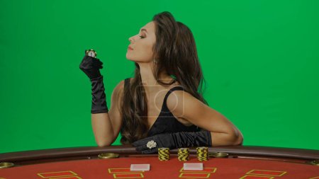 Photo for Casino and gambling commercial advertisement concept. Female in studio on chroma key green screen close up. Woman in black dress sitting at the blackjack poker table smiling holding chip, side view. - Royalty Free Image