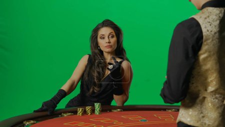 Photo for Casino and gambling commercial advertisement concept. Gorgeous female in studio on chroma key green screen. Appealing woman in dress and man croupier at the blackjack poker table looks at cards. - Royalty Free Image