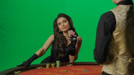 Photo for Casino and gambling commercial advertisement concept. Gorgeous female in studio on chroma key green screen. Appealing woman in dress and man croupier at the blackjack poker table looks at man. - Royalty Free Image