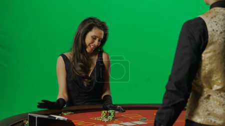 Photo for Casino and gambling commercial advertisement concept. Gorgeous female in studio on chroma key green screen. Appealing woman in dress and man croupier at the blackjack poker table happy expression. - Royalty Free Image