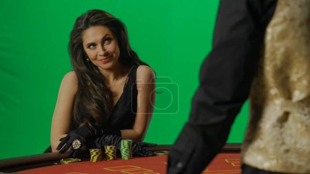 Photo for Casino and gambling commercial advertisement concept. Gorgeous female in studio on chroma key green screen. Appealing woman in dress and man croupier at the blackjack poker table smiling at him. - Royalty Free Image