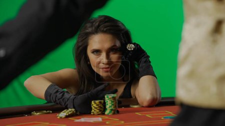 Photo for Casino and gambling commercial advertisement concept. Gorgeous female in studio on chroma key green screen. Appealing woman in dress and man croupier at the blackjack poker table looks at camera. - Royalty Free Image