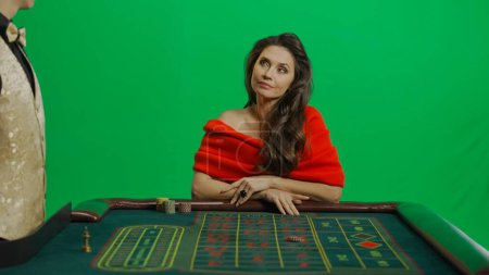 Photo for Casino and gambling commercial advertisement concept. Gorgeous female in studio on chroma key green screen. Appealing woman in red dress and male croupier at the roulette table looks at man. - Royalty Free Image