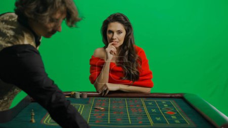 Photo for Casino and gambling commercial advertisement concept. Gorgeous female in studio on chroma key green screen. Appealing woman in red dress and male croupier at the roulette table placing bet thinking. - Royalty Free Image