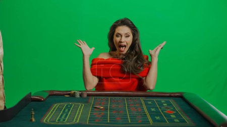 Photo for Casino and gambling commercial advertisement concept. Gorgeous female in studio on chroma key green screen. Appealing woman in red dress sitting at the roulette table won the game screaming happily. - Royalty Free Image
