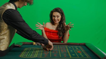 Photo for Casino and gambling commercial advertisement concept. Gorgeous female in studio on chroma key green screen. Appealing woman in red dress and male croupier at the roulette table takes winning money. - Royalty Free Image