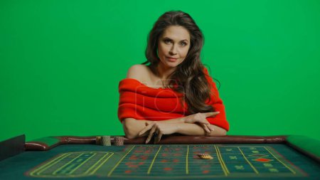 Photo for Casino and gambling commercial advertisement concept. Gorgeous female in studio on chroma key green screen. Appealing woman in red dress sitting at the roulette table looking directly at camera. - Royalty Free Image