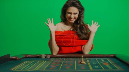 Photo for Casino and gambling commercial advertisement concept. Gorgeous female in studio on chroma key green screen. Appealing woman in red dress sitting at the roulette table smiling happy winner gesture. - Royalty Free Image