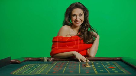 Photo for Casino and gambling commercial advertisement concept. Gorgeous female in studio on chroma key green screen. Appealing woman in red dress sitting at the roulette table smiling widely at camera. - Royalty Free Image