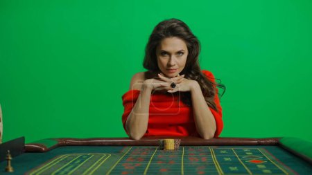 Photo for Casino and gambling commercial advertisement concept. Gorgeous female in studio on chroma key green screen. Appealing woman in red dress sitting at the roulette table looks serious at camera. - Royalty Free Image