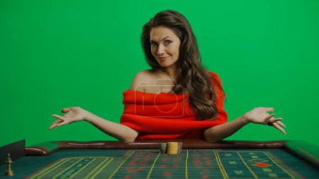Photo for Casino and gambling commercial advertisement concept. Gorgeous female in studio on chroma key green screen. Appealing woman in red dress sitting at the roulette table smiling with spread hands. - Royalty Free Image