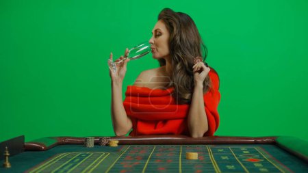 Photo for Casino and gambling commercial advertisement concept. Gorgeous female in studio on chroma key green screen. Appealing woman in red dress sitting at the roulette table drinks glass of champagne. - Royalty Free Image