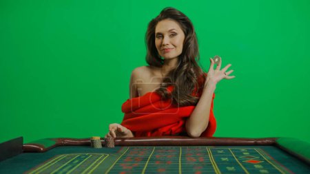 Photo for Casino and gambling commercial advertisement concept. Gorgeous female in studio on chroma key green screen. Appealing woman in red dress sitting at the roulette table smiling holding chip. - Royalty Free Image