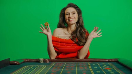 Photo for Casino and gambling commercial advertisement concept. Gorgeous female in studio on chroma key green screen. Appealing woman in red dress sitting at the roulette table laughs holding chips. - Royalty Free Image
