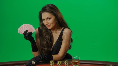 Photo for Casino and gambling commercial advertisement concept. Female in studio on chroma key green screen isolated background. Woman in black dress at the blackjack poker table holds cards, smiles posing. - Royalty Free Image