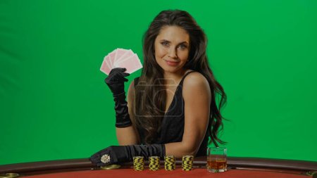 Photo for Casino and gambling commercial advertisement concept. Female in studio on chroma key green screen isolated background. Woman in black dress at the blackjack table posing with whiskey and cards. - Royalty Free Image