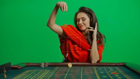 Photo for Casino and gambling commercial advertisement concept. Gorgeous female in studio on chroma key green screen. Appealing woman in red dress at the roulette table at camera drops chip on the table. - Royalty Free Image