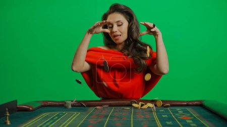 Photo for Casino and gambling commercial advertisement concept. Gorgeous female in studio on chroma key green screen. Appealing woman in red dress at the roulette table dropping many chips on the table. - Royalty Free Image