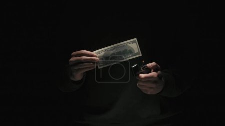 Foto de Concepto de publicidad creativa de negocios y dinero. Retrato de hombre en ropa negra aislado sobre fondo negro con poca luz. Hombre sosteniendo billetes de cien dólares y encendiendo el encendedor - Imagen libre de derechos