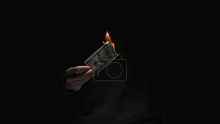 Photo for Business and money creative advertisement concept. Portrait of male in black clothes isolated on black background in low light. Man burns hundred dollar banknote - Royalty Free Image