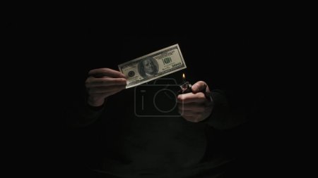 Foto de Concepto de publicidad creativa de negocios y dinero. Retrato de hombre en ropa negra aislado sobre fondo negro con poca luz. Hombre sosteniendo billetes de cien dólares y encendiendo el encendedor - Imagen libre de derechos