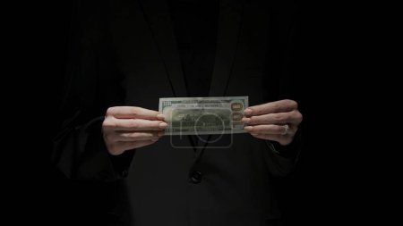 Photo for Business and money creative advertisement concept. Portrait of female in black suit isolated on black background in low light. Woman hands close up holding a dollar banknote backside at the camera. - Royalty Free Image