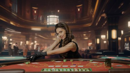 Foto de Atractiva mujer en vestido negro en la mesa de póquer para el juego de blackjack en el casino. Una mujer mira a la cámara y sonríe, hay cartas y fichas en la mesa. Concepto de casino y juegos de azar - Imagen libre de derechos