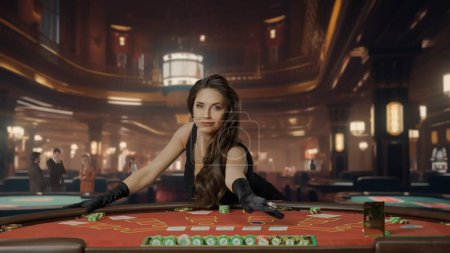 Téléchargez les photos : Belle femme en robe noire à la table de poker pour le jeu de blackjack au casino. Une femme regarde dans la caméra et sourit, il y a des cartes et des jetons sur la table. Concept de casino et de jeu - en image libre de droit