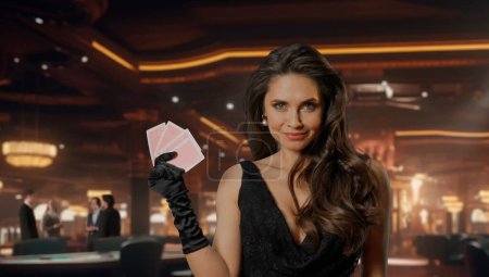 Téléchargez les photos : Femme chic en robe noire à la table de poker pour le jeu de blackjack au casino. Femme tenant des cartes de poker, regardant la caméra et souriant. Le concept de casino et de jeu - en image libre de droit