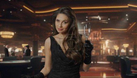 Téléchargez les photos : Femme chic en robe noire à la table de poker pour le jeu de blackjack au casino. La femme tient un verre de champagne dans sa main et sourit à la caméra. Le concept de casino et de jeu - en image libre de droit