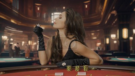 Téléchargez les photos : Femme chic en robe noire à la table de poker pour le jeu de blackjack au casino. La femme regarde une puce d'une valeur de cent dollars. Le concept de casino et de jeu - en image libre de droit