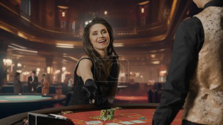 Téléchargez les photos : Belle femme en robe noire à la table de poker pour le jeu de blackjack au casino. La femme sourit joyeusement et tend la main pour ses jetons gagnants. Le concept de casino et de jeu - en image libre de droit
