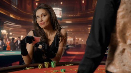 Foto de Atractiva mujer en vestido negro en la mesa de póquer para el juego de blackjack en el casino. Mujer sosteniendo cartas de póquer y mirando pensativamente al crupier. Concepto de casino y juegos de azar - Imagen libre de derechos