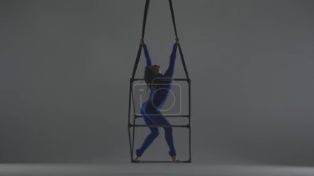 Téléchargez les photos : Chorégraphie moderne et acrobaties concept publicitaire créatif. Portrait d'acrobate femelle isolé sur fond gris. Femme danseuse aerialiste en costume bleu filant en l'air sur cube avec cordes. - en image libre de droit