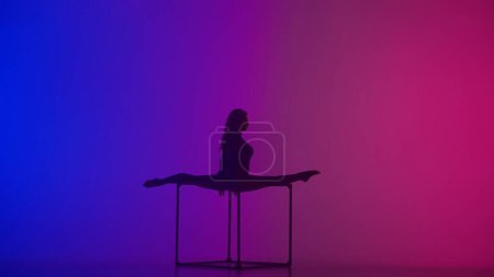 Photo for Modern choreography and acrobatics creative advertisement concept. Silhouette of female acrobat isolated on neon background. Woman gymnastic dancer in blue sparkling bodysuit showing split on a cube. - Royalty Free Image