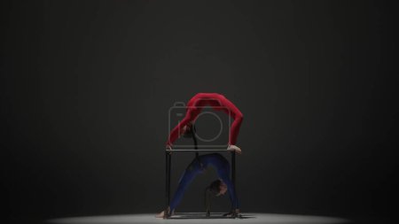 Téléchargez les photos : Chorégraphie moderne et acrobaties concept publicitaire créatif. Portrait de deux acrobates femelles isolées sur fond noir. Filles danseuses aériennes en costumes bleus rouges effectuant pont sur un cube. - en image libre de droit