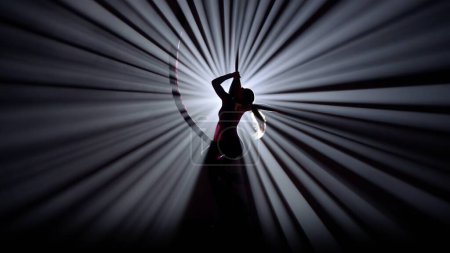 Modern choreography and acrobatics creative advertisement concept. Silhouette of female isolated on black background against bright spotlight. Girl aerial dancer performs element on air silk.