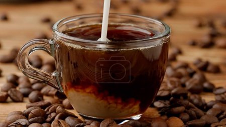 Téléchargez les photos : Un jet de lait se déverse dans une tasse pleine de café et s'y dissout. Gros plan d'une tasse de café ou de thé en verre sur la table de cuisine. Un rituel matinal, un concept de petit déjeuner - en image libre de droit